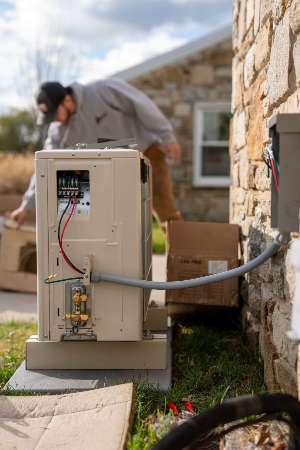 A person performs a mini split install San Diego next to a stone building, with the outdoor HVAC unit's wiring and components exposed and cardboard boxes scattered nearby on the ground.