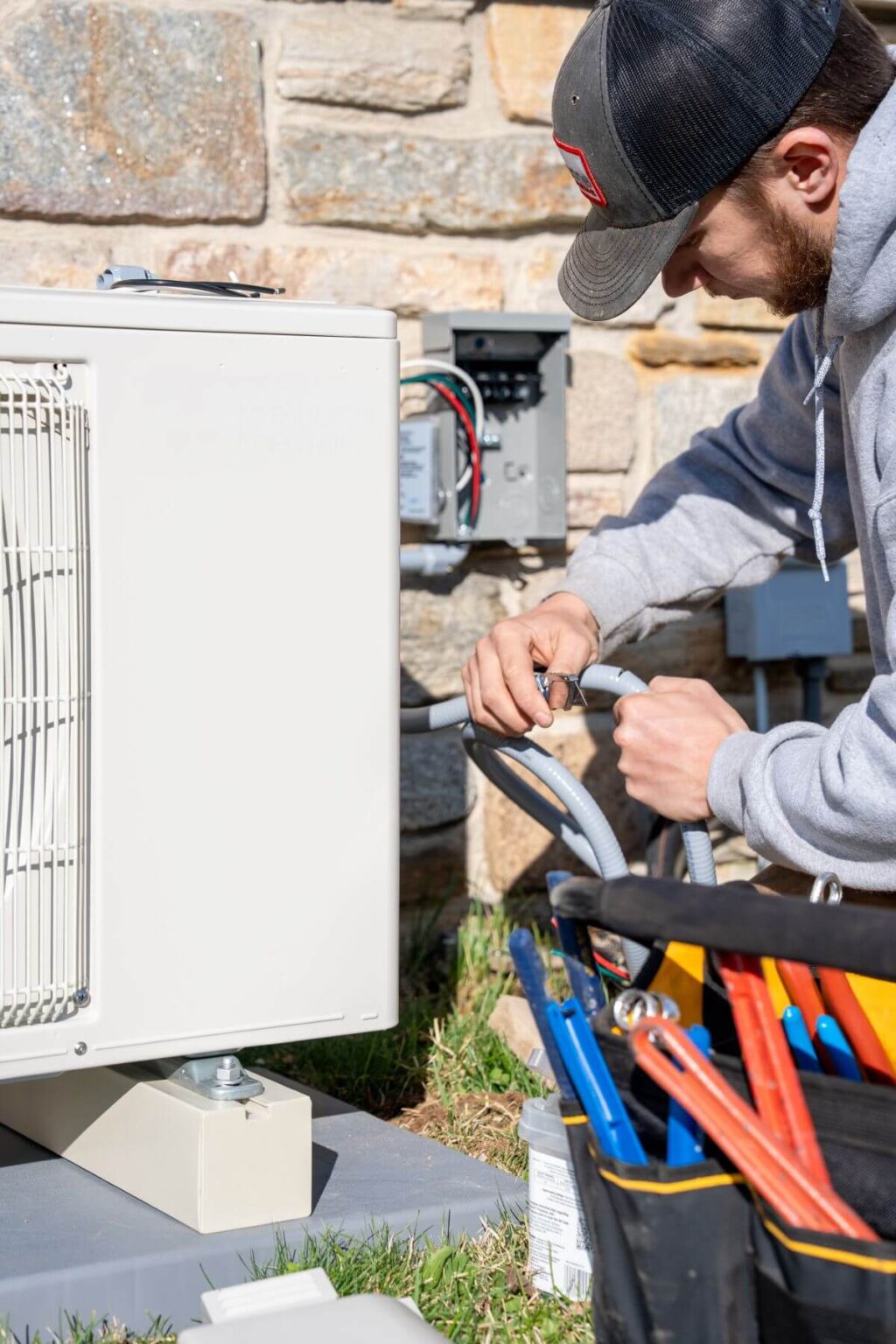 A person wearing a gray hoodie and black cap works on wiring an outdoor HVAC unit during a mini split install San Diego near a stone wall. A tool bag with various tools is visible in the foreground on grass.