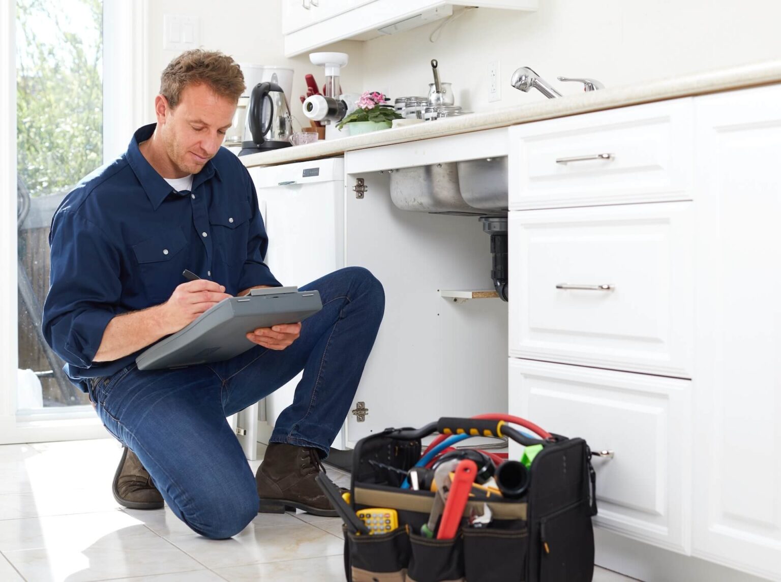 A person in a blue shirt kneels by an open cabinet under a kitchen sink, writing on a clipboard during a Plumbing Inspection San Diego. A tool bag with various tools sits nearby in the kitchen with white cabinets and countertops.