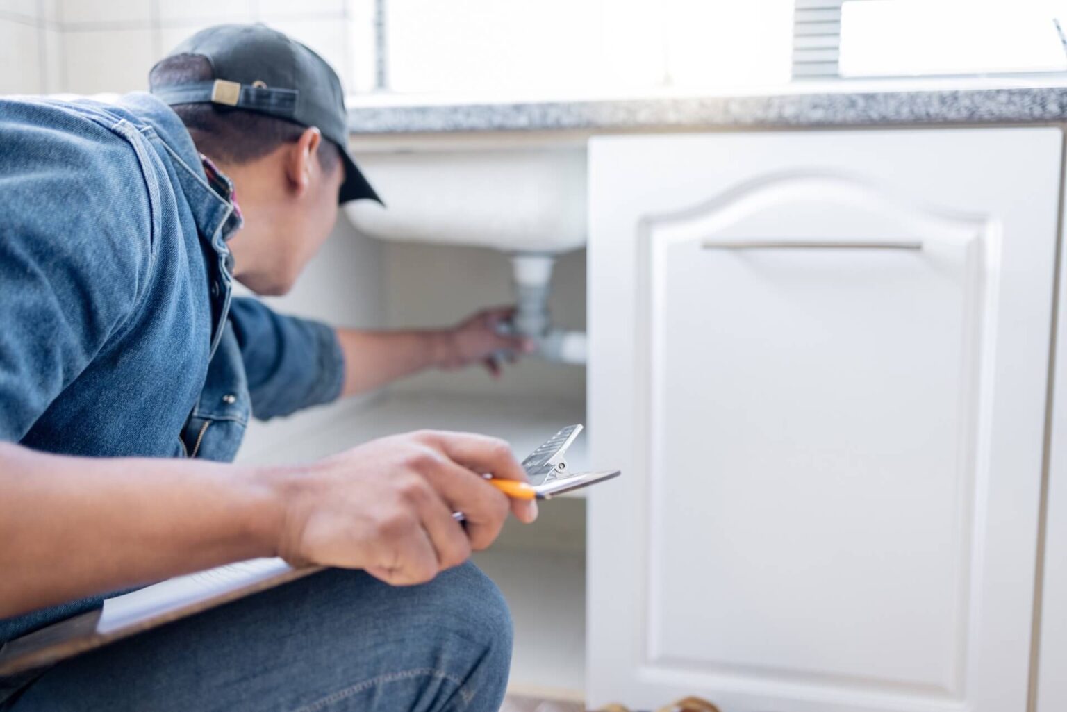 A person wearing a denim shirt and cap kneels in front of a kitchen sink cabinet, holding pliers and inspecting pipes under the sink—a clear example of a thorough Plumbing Inspection San Diego. The focus is on their actions rather than their face.