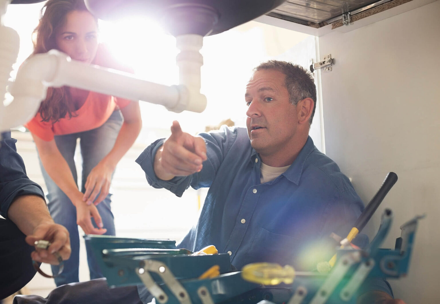 A man lies under a sink pointing at pipes while a woman leans in to watch. Various tools are visible in the foreground during a plumbing repair in Fallbrook, CA, as sunlight shines through the background.