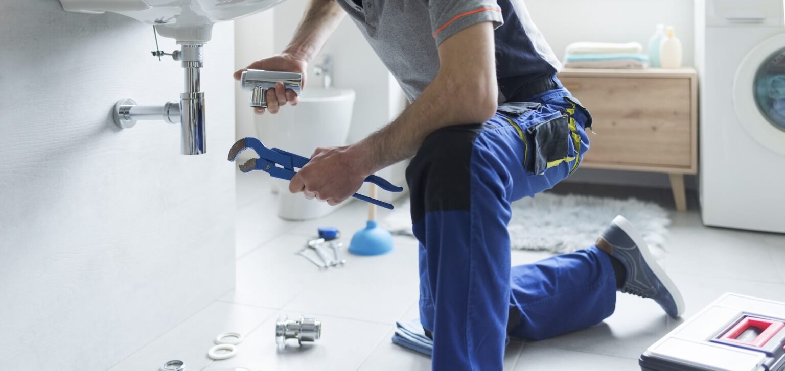 A person in work clothes kneels beside a bathroom sink, using a wrench to repair the plumbing—possibly part of a Repiping Installation San Diego project. Various tools and parts are scattered nearby, with a washing machine in the background.