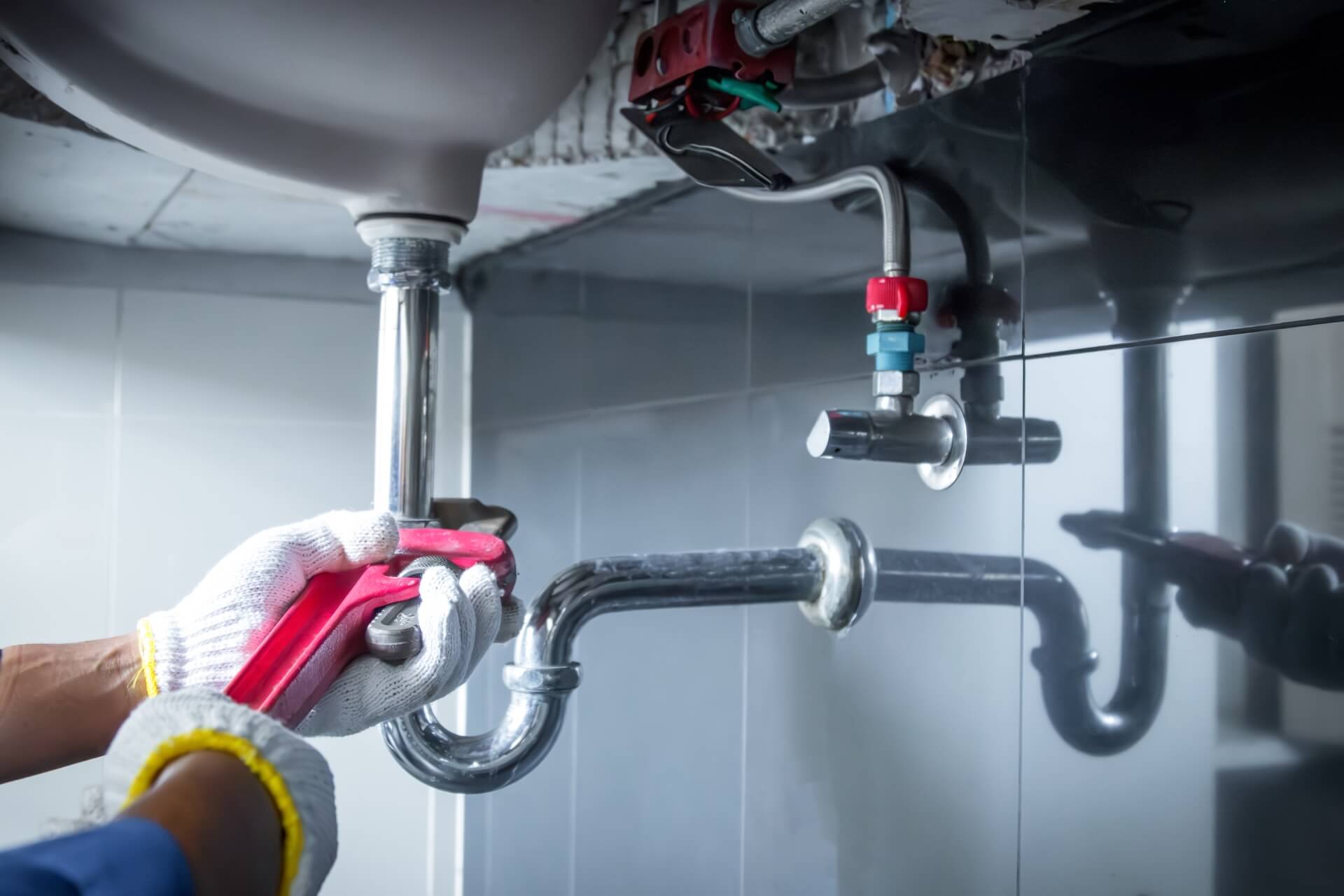 A person wearing white work gloves uses a red pipe wrench to tighten or loosen plumbing pipes under a sink, showcasing expert Repiping Installation San Diego in a bathroom or kitchen.