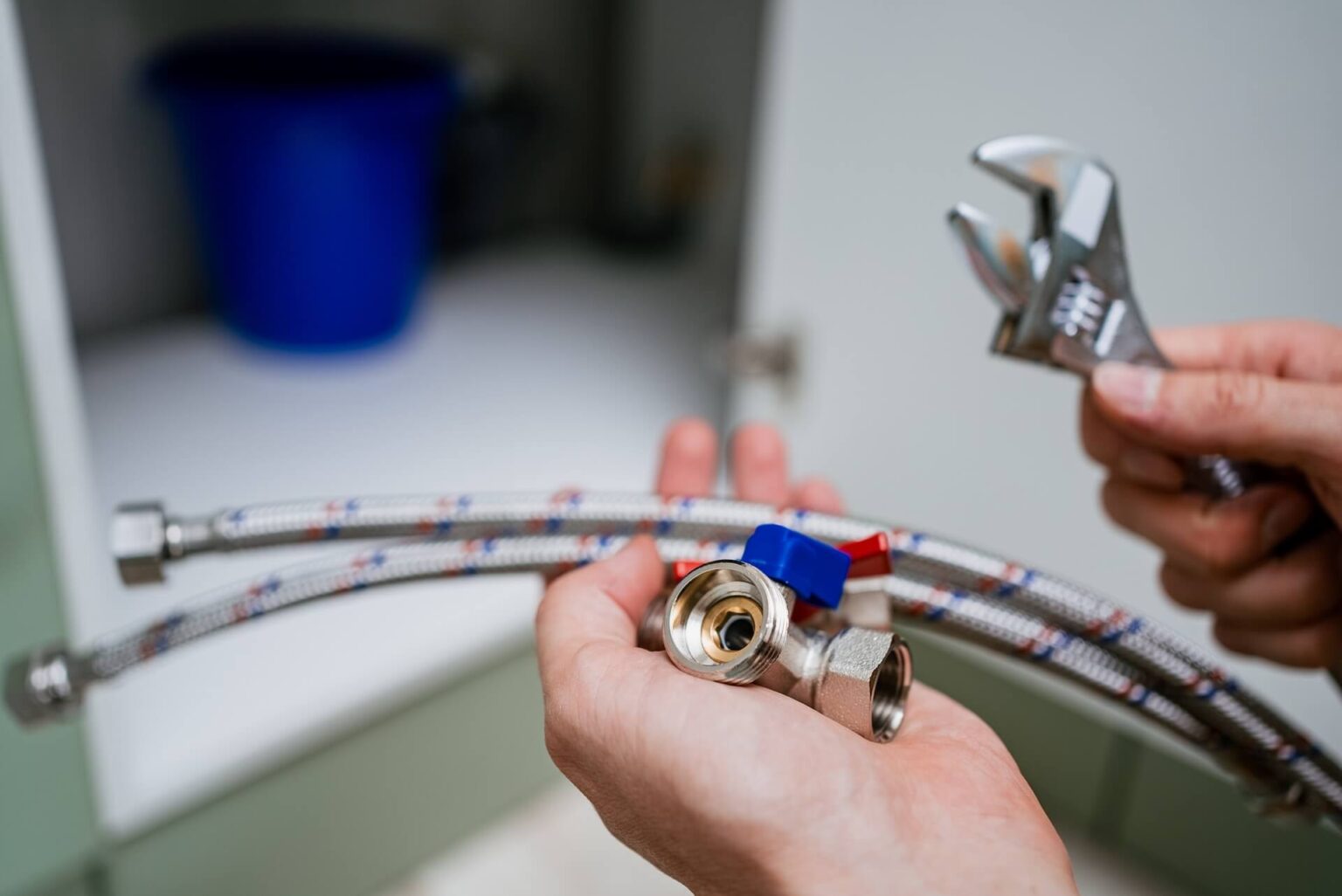 A person holds flexible water supply hoses, a plumbing fitting, and a wrench, preparing for Repiping Installation San Diego projects. A blue bucket is visible in the background under a sink.