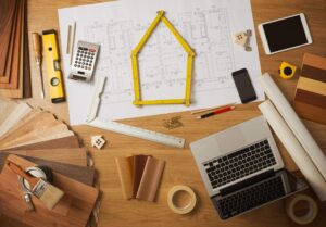 A desk with architectural plans, a laptop, wood samples, measuring tools, a calculator, a smartphone, a tablet, tape, pencils, and a folded ruler arranged in the shape of a house.
