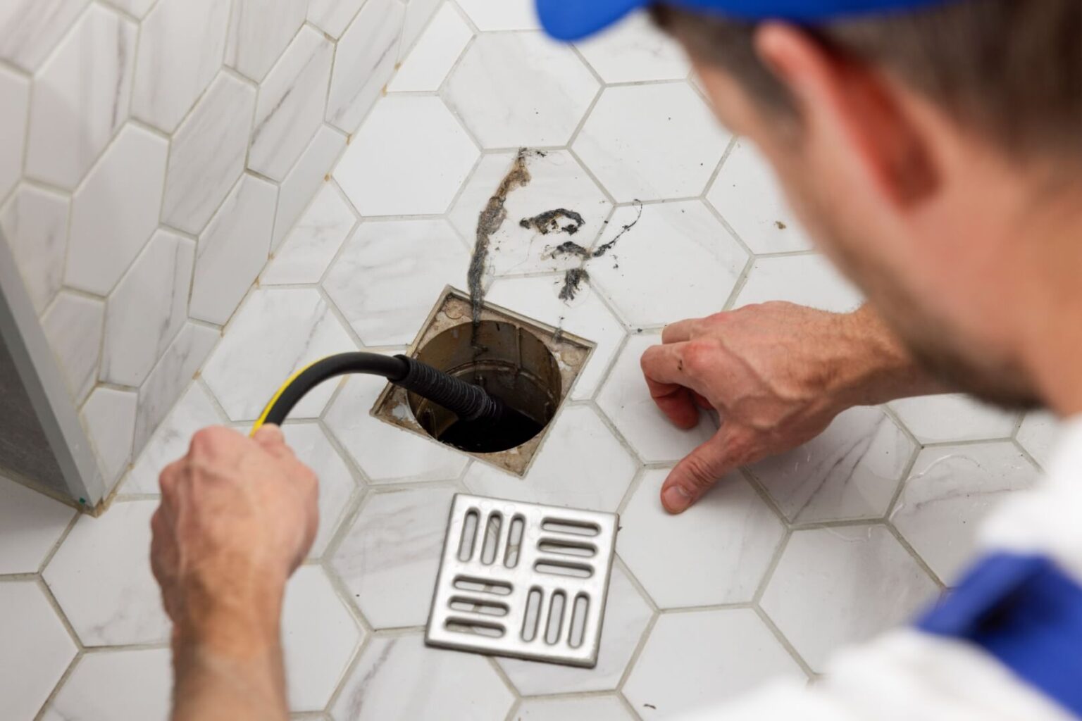 A person in work clothes uses a flexible tool to clean or inspect a square floor drain on white hexagonal tile, with the metal drain cover removed and set nearby—demonstrating expert Rooter Services San Diego.