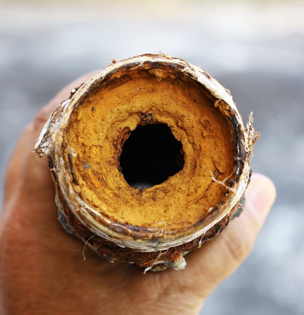 A hand holds a corroded metal pipe with thick, orange-brown mineral buildup and a partially blocked central hole, highlighting severe internal clogging—a common issue solved by Rooter Services San Diego.