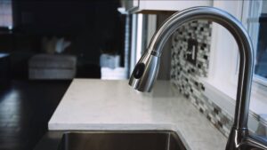 A close-up of a modern stainless steel kitchen faucet over a white marble countertop with mosaic tile backsplash; a sink and a partially visible living room are in the background.