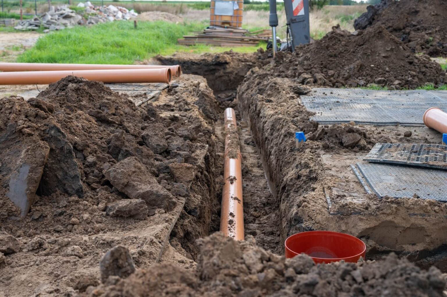 Orange plastic pipes are laid in a trench at a sewer line install San Diego site, surrounded by piles of soil, a red bucket, and metal grates. Grass and equipment are visible in the background at this active construction area.