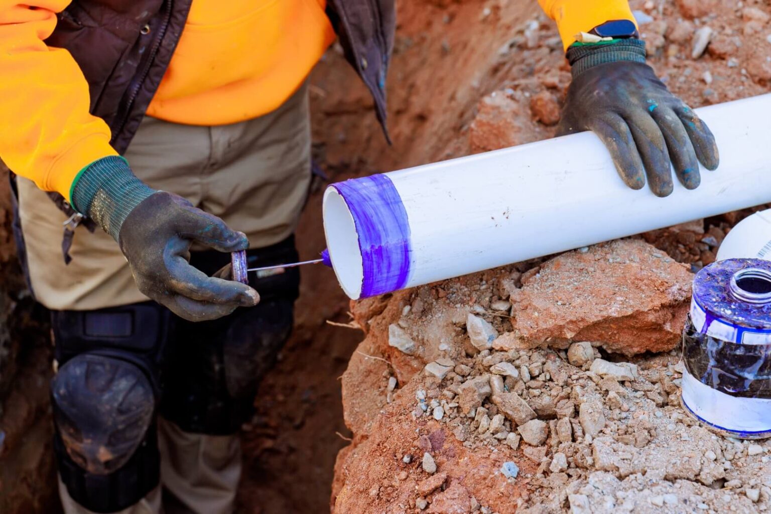 A person wearing gloves and a yellow jacket applies purple primer to the end of a white PVC pipe during a sewer line install San Diego project, with dirt and rocks visible on the construction site ground.