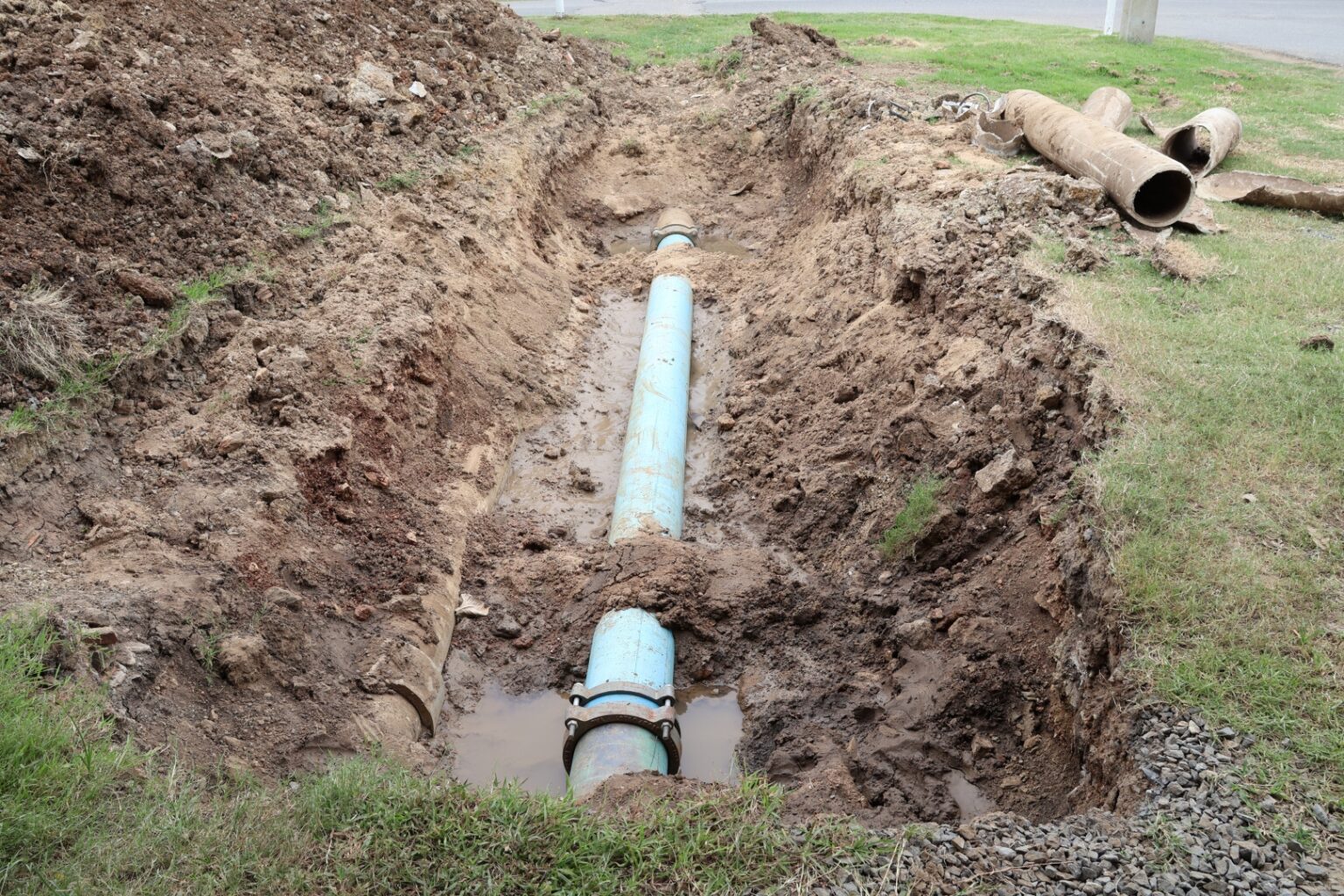 A large blue pipe is partially buried in a trench surrounded by soil and grass, with some water collecting near the pipe—a common scene during sewer line repair San Diego homeowners might encounter. Sections of removed pipe and dirt are visible nearby.