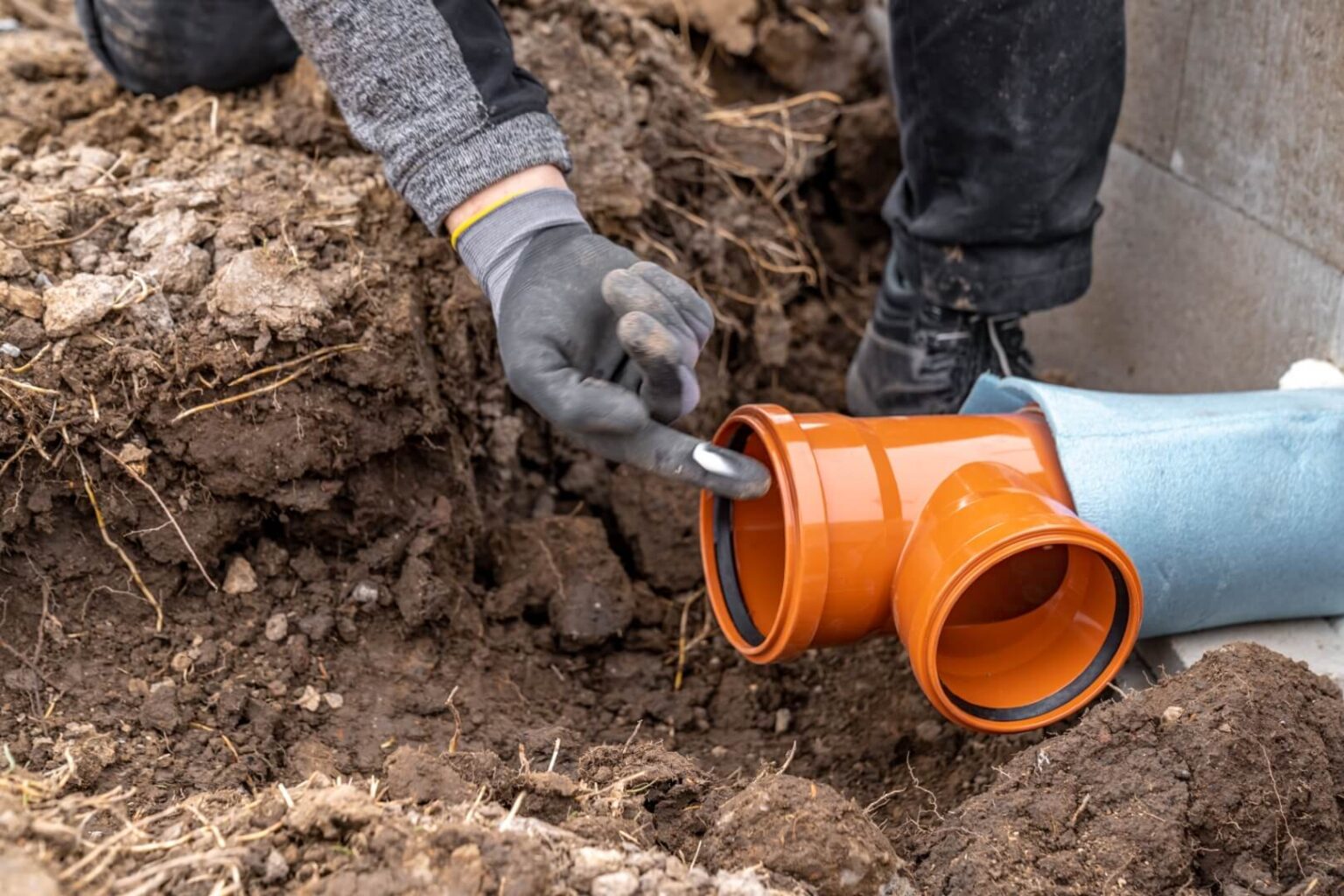 A person wearing gloves is connecting two orange PVC pipes in a trench surrounded by soil. One hand applies a substance, possibly lubricant or sealant, to the pipe’s end—a typical step in sewer line repair San Diego professionals perform.