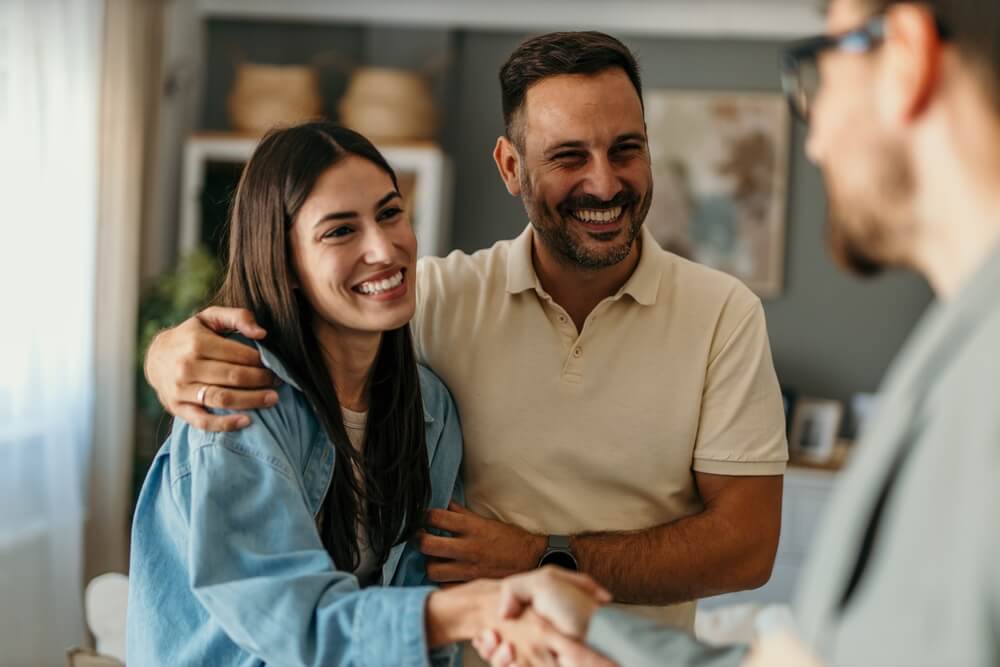 A smiling man with an arm around a smiling woman stands indoors while shaking hands with another person whose face is partially visible in the foreground.