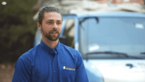 A man with dark hair pulled back, wearing a blue collared shirt with a logo, stands outdoors in front of a blue vehicle.