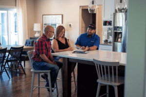 Three people sit around a kitchen island; a man in a blue shirt and cap shows documents to a man in a red plaid shirt and a woman in black. They appear to be discussing paperwork in a modern home setting.