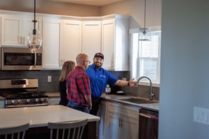 A man in a blue uniform speaks to a man and woman in a kitchen, gesturing toward the sink area. The kitchen has white cabinets, a stove, a microwave, and a pendant light over a counter.