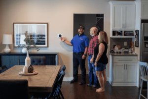 A technician in a blue shirt shows a wall-mounted thermostat to a man and woman in a kitchen and dining area, as they all look at the device and smile.