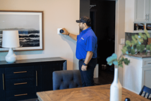 A person in a blue shirt and cap adjusts a thermostat on a wall in a modern living space with a dining table, a lamp, a framed picture, and a vase on a sideboard.