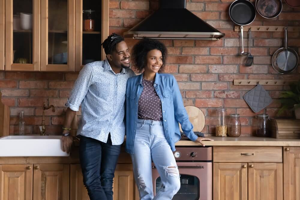 A man and woman stand together in a modern kitchen with wood cabinets and a brick backsplash, smiling as they enjoy the perks of their Sherlock Smart Membership. Various kitchen items and utensils hang on the wall behind them.