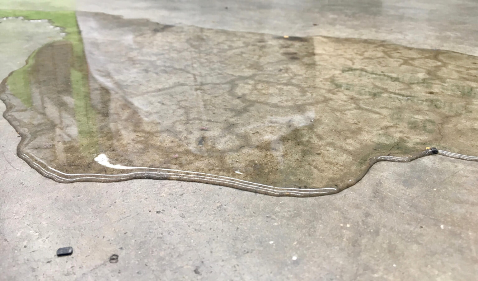 A close-up view of a puddle of clear liquid, possibly from a slab leak service San Diego homeowners might need, spreading across a rough, light gray concrete surface. The liquid has an irregular shape and reflects some light.