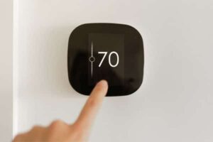 A hand is adjusting a black digital thermostat mounted on a white wall, which displays the temperature set to 70 degrees.
