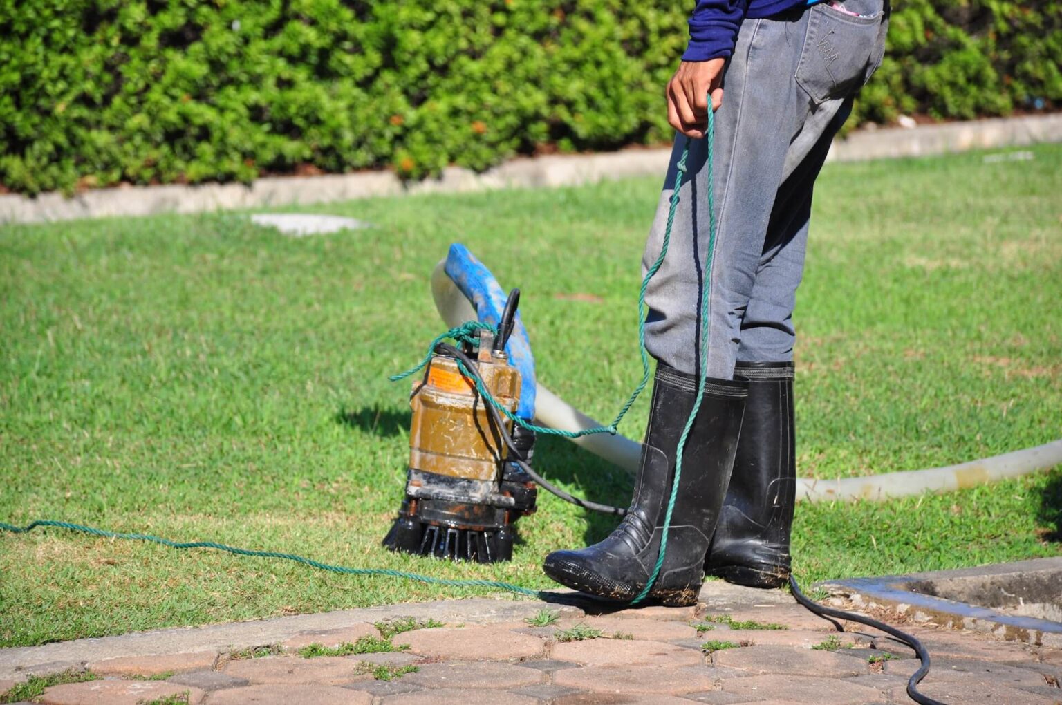 A person wearing black rubber boots and gray pants stands on grass holding a rope attached to a sump pump install San Diego setup, with a hose connected, near a paved area and green shrubs in the background.