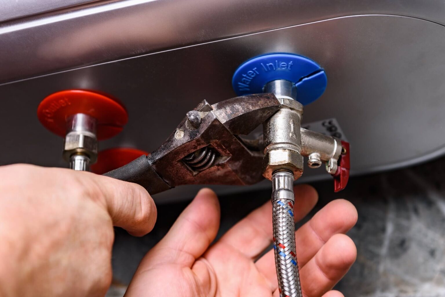 A hand uses a wrench to tighten a metal hose connected to the blue water inlet valve on a water heater during a Tankless Water Heater Install San Diego. A red hot water valve is also visible next to the blue valve.
