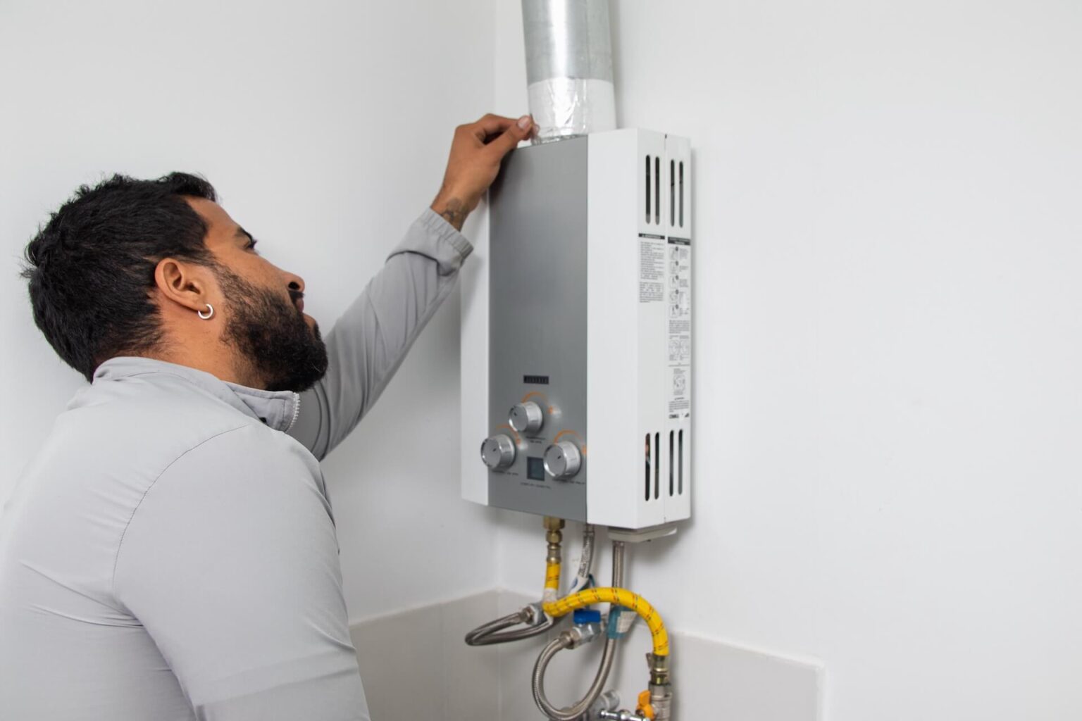 A person inspects a wall-mounted gas water heater with visible pipes and a vent, inside a clean, white-walled room—typical of a professional Tankless Water Heater Install San Diego homeowners rely on.