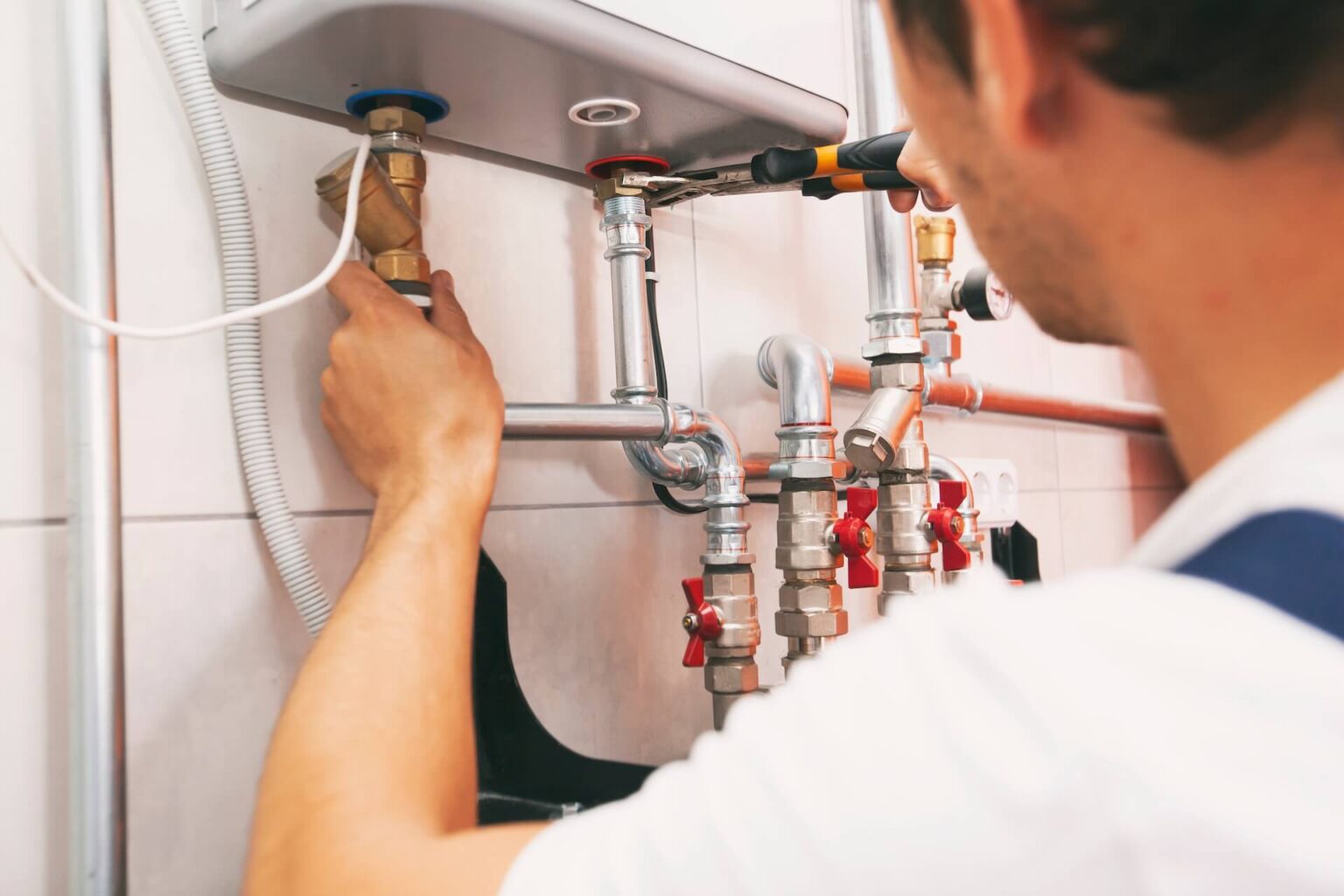 A plumber adjusts pipes and valves beneath a tankless water heater, using a wrench, with various metal pipes and fittings visible against a tiled wall—perfect for precise tankless water heater repair.