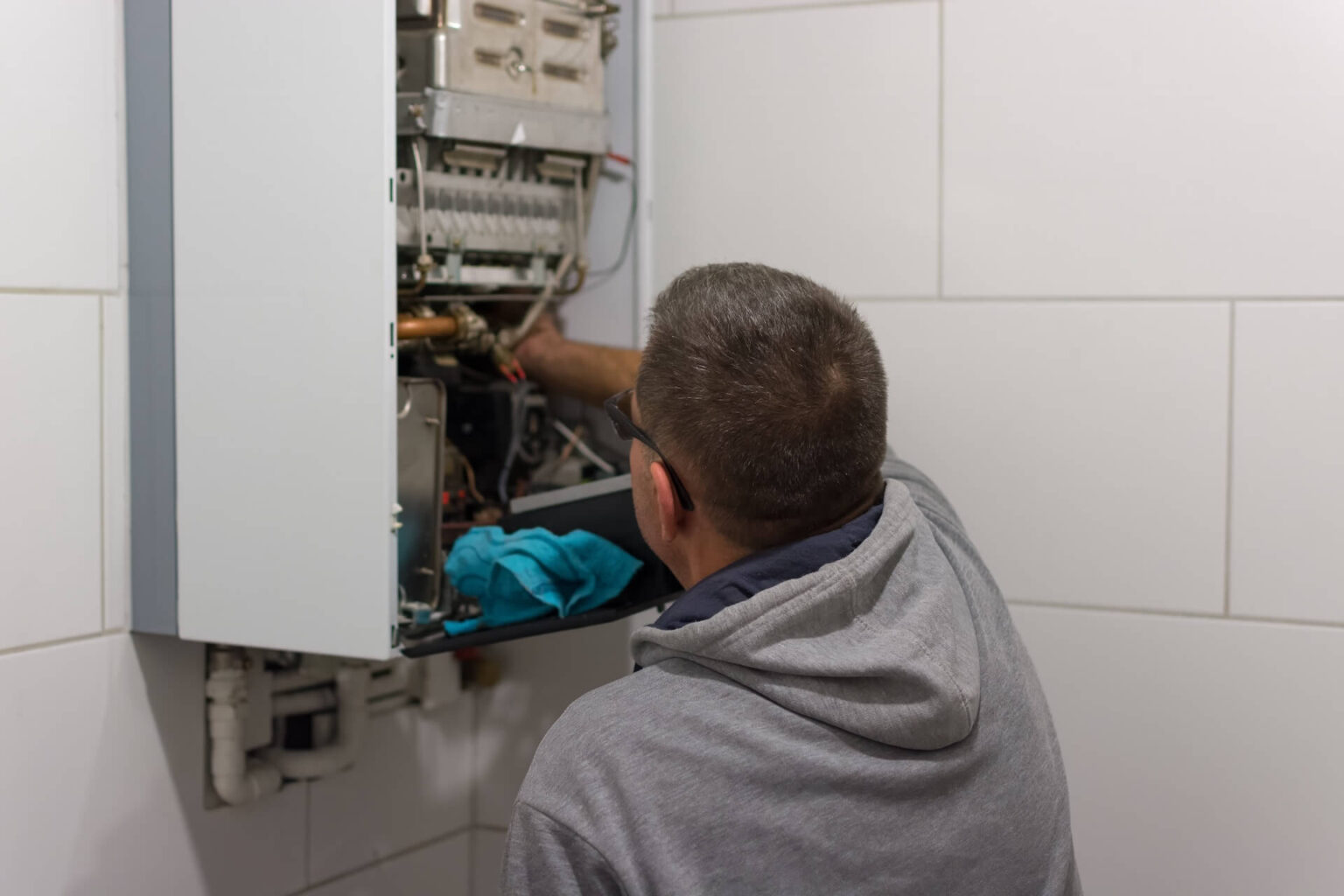 A person wearing glasses and a gray hoodie is working on a tankless water heater mounted on a white tiled wall, with tools and a blue cloth nearby, focusing on precise tankless water heater repair.