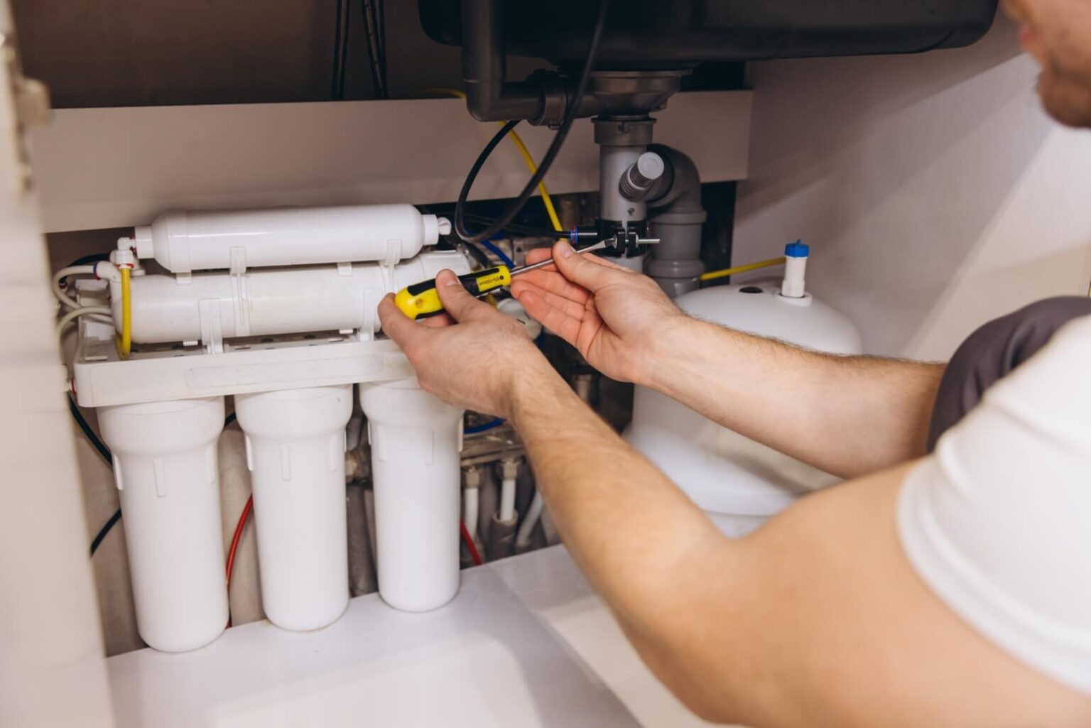 A person uses a screwdriver to work on a water filtration installation in San Diego under a sink. Several cylindrical filter cartridges and pipes are visible in the system.