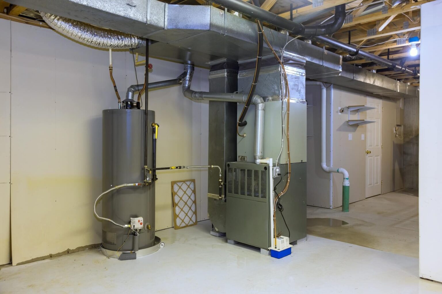 A basement utility room features Water Heater Installation San Diego, a furnace, exposed ducts and pipes, an air filter, and an unfinished ceiling. The walls are partially finished, with a closed door visible in the background.