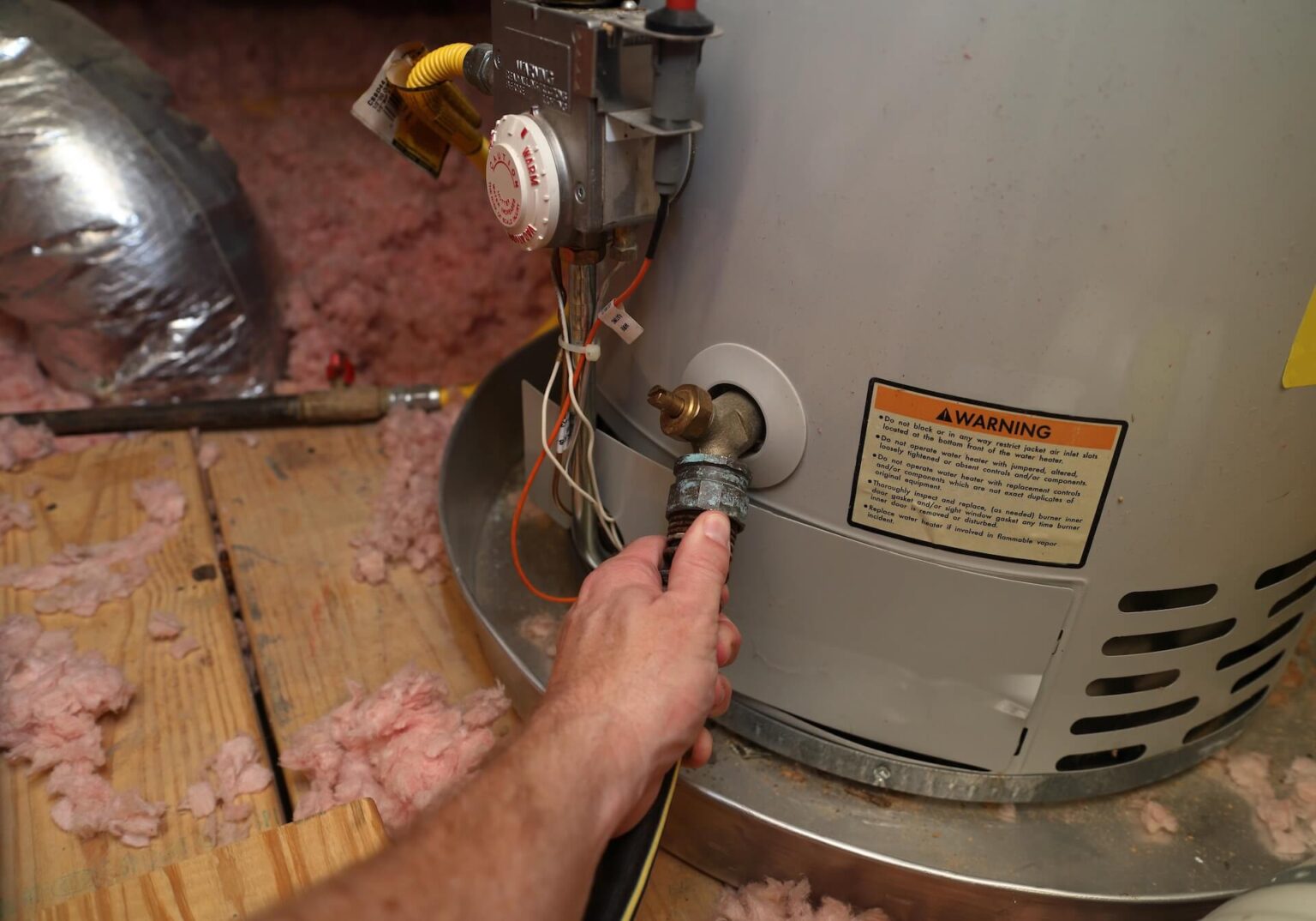 A person’s hand is attaching a hose to the drain valve at the bottom of a water heater tank in an attic or utility space, highlighting essential Water Heater Maintenance San Diego residents can trust.