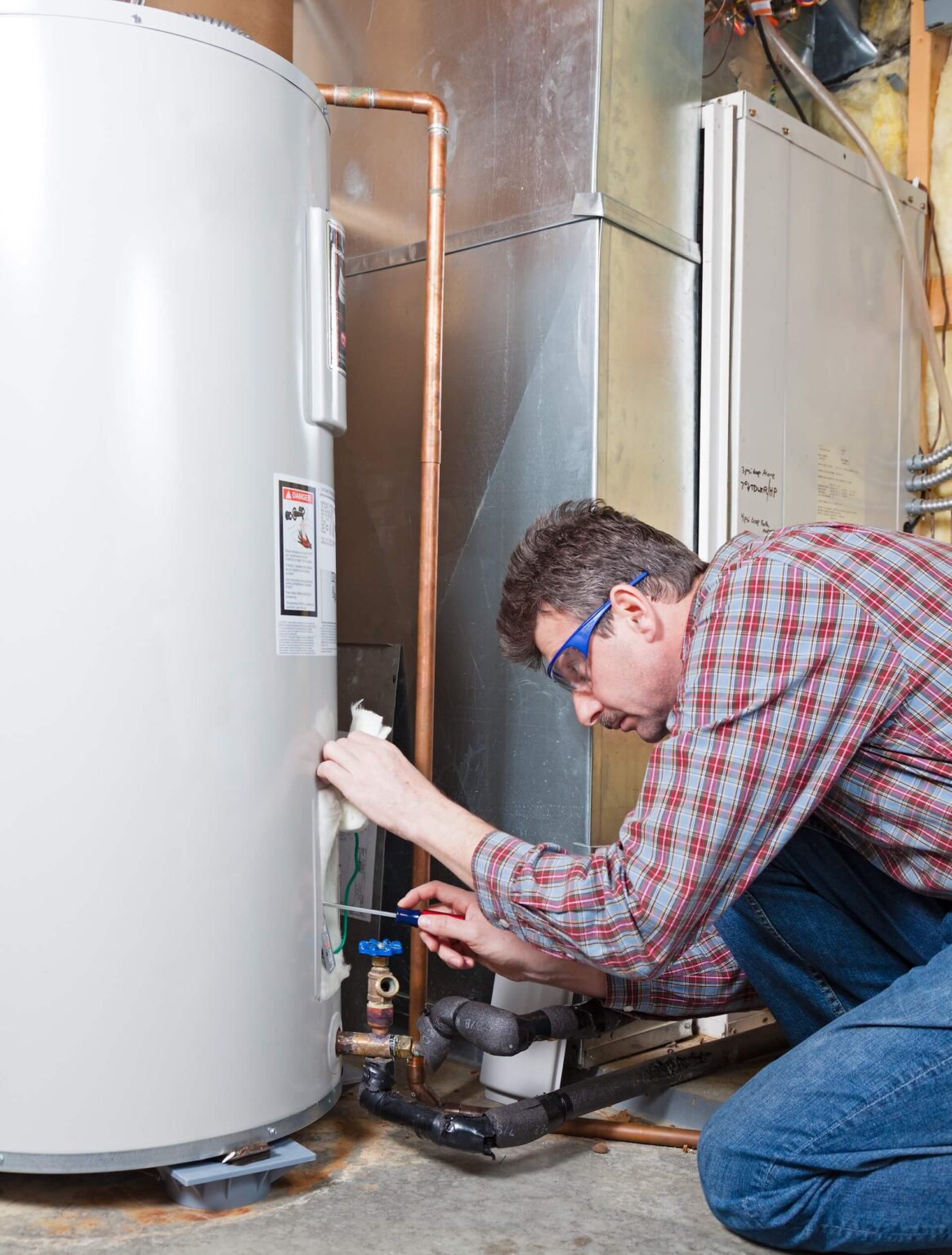A man wearing glasses and a plaid shirt kneels next to a water heater in a basement, using a tool to adjust or repair a pipe—showcasing expert Water Heater Repair San Diego services.