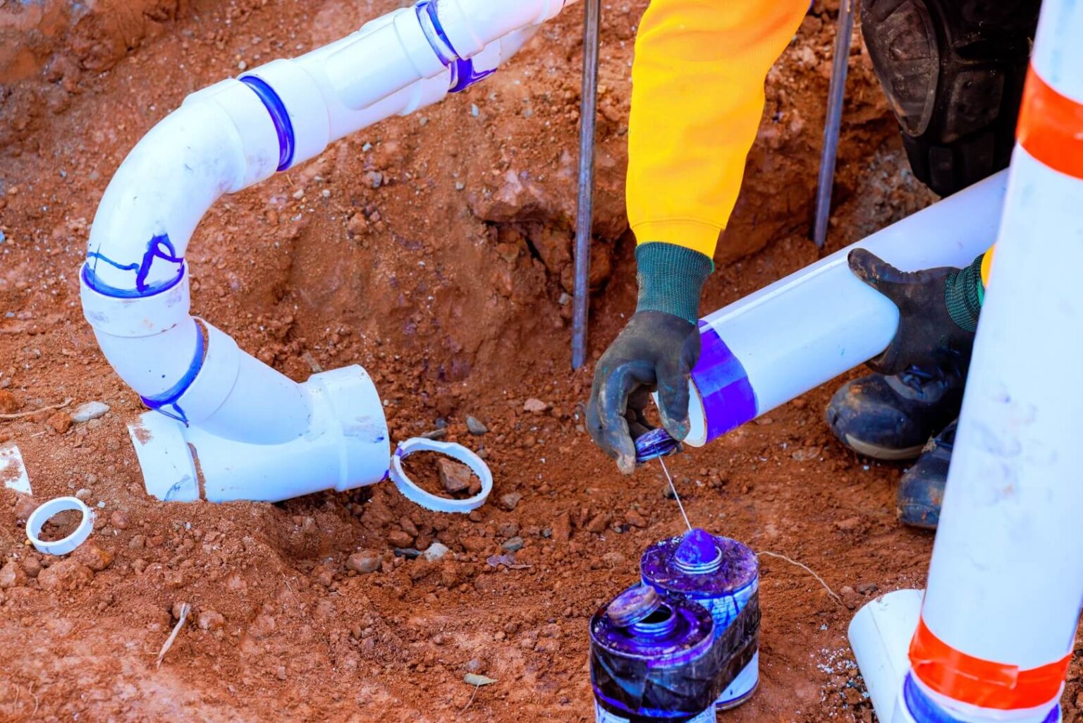 A person wearing gloves and a yellow shirt applies purple PVC primer to a white pipe joint during a water line install San Diego project at a construction site with exposed soil and other pipes nearby.