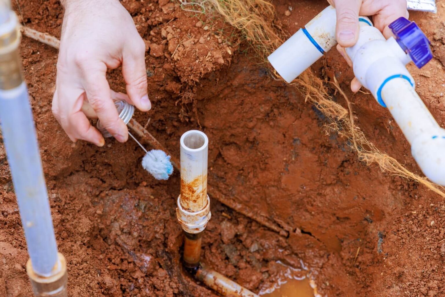 A person applies pipe glue to a white PVC pipe outdoors, preparing to join it with another for a water line install San Diego residents trust. The pipes are set in a shallow trench with exposed soil and mud visible around the plumbing work.