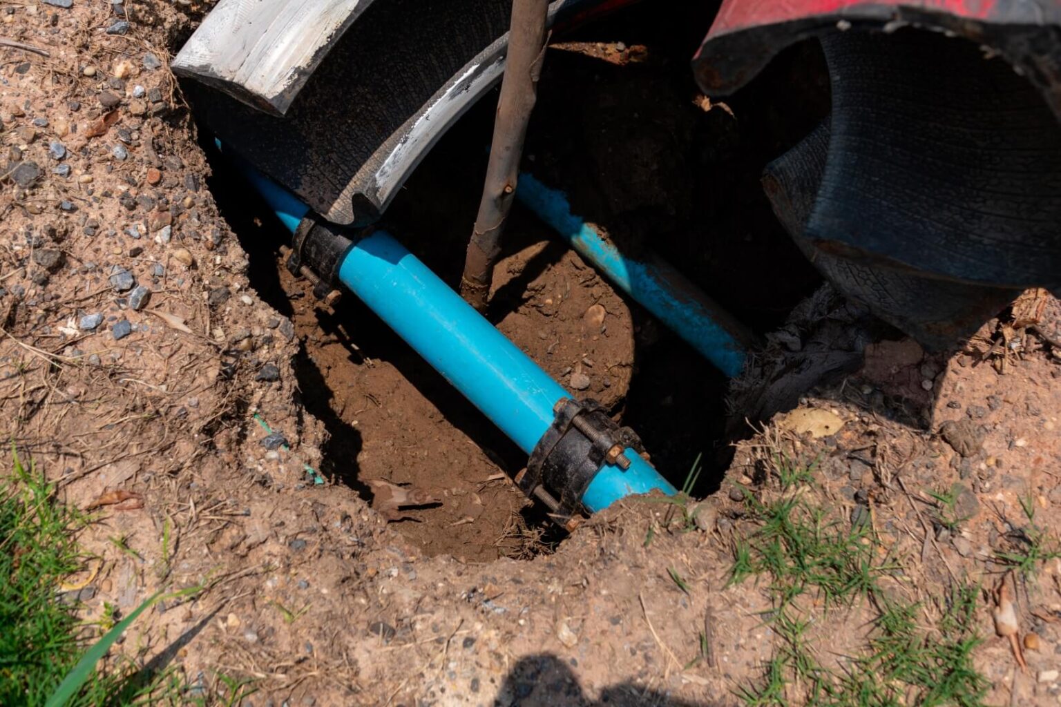 A blue underground water pipe exposed in a dirt hole, with black pipe fittings and protective covering pulled back—typical of a Water Line Repair San Diego project. Grass and scattered gravel border the excavation.