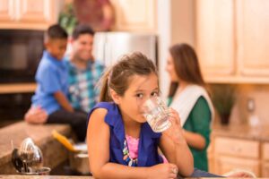 A young girl drinks a glass of water at a kitchen counter while a boy and two adults talk in the background. The kitchen has wooden cabinets and a granite countertop.