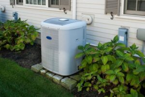 A central air conditioning unit is installed outside a house next to a garden bed with green plants, sitting on a stone base against the exterior siding wall.