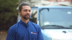 A man with a beard and tied-back hair wearing a blue button-up shirt stands in front of a blue and white van. The van has equipment on its roof, and trees are visible in the background.