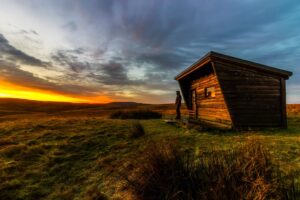 A small wooden cabin stands on grassy terrain under a dramatic, cloudy sky during sunset, with warm orange light illuminating the horizon in the distance.
