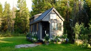 A small, dark gray wooden tiny house with multiple windows sits on a grassy lawn surrounded by trees and garden planters, with sunlight filtering through the surrounding forest.